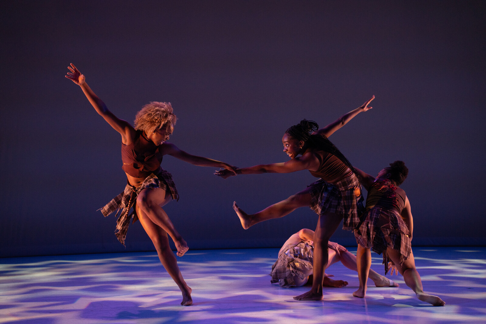 Four dancers perform on stage under purple-blue lighting, capturing a dynamic moment of physical interaction with expressive poses. The setting suggests a contemporary dance performance.