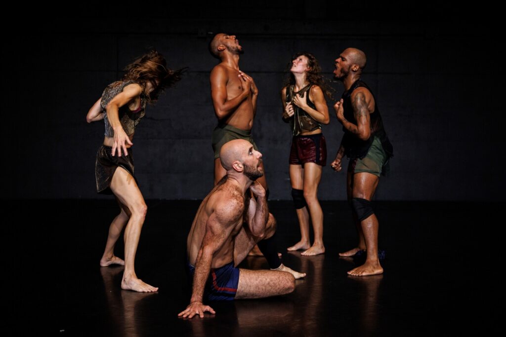 Five dancers posed on a dark stage in different expressions of anguish and relief